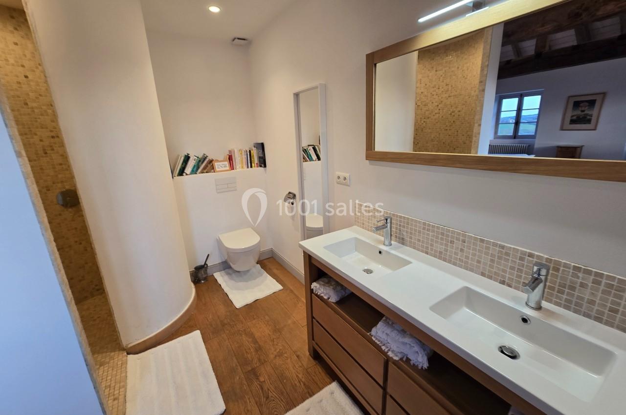 Salle de bain avec double vasque, douche à l'italienne, toilettes, étagères avec livres et sol en bois.