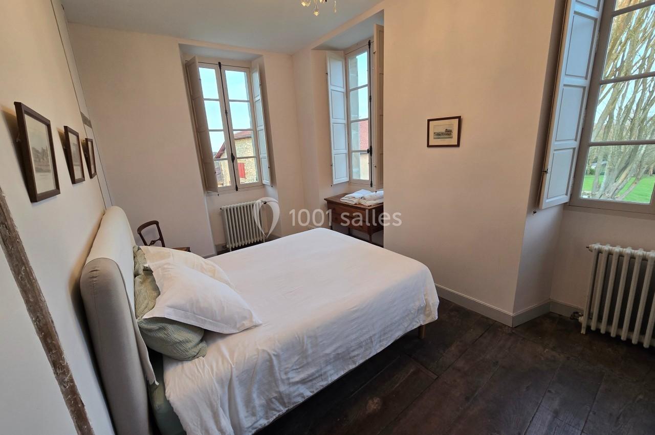 Chambre lumineuse avec lit double, parquet en bois sombre, murs clairs, fenêtres ouvertes et mobilier en bois.