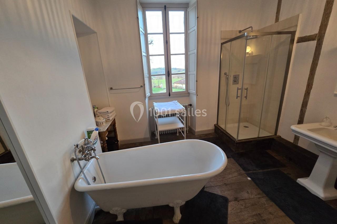 Salle de bain avec baignoire sur pieds, douche vitrée, lavabo et fenêtre donnant sur un paysage champêtre.