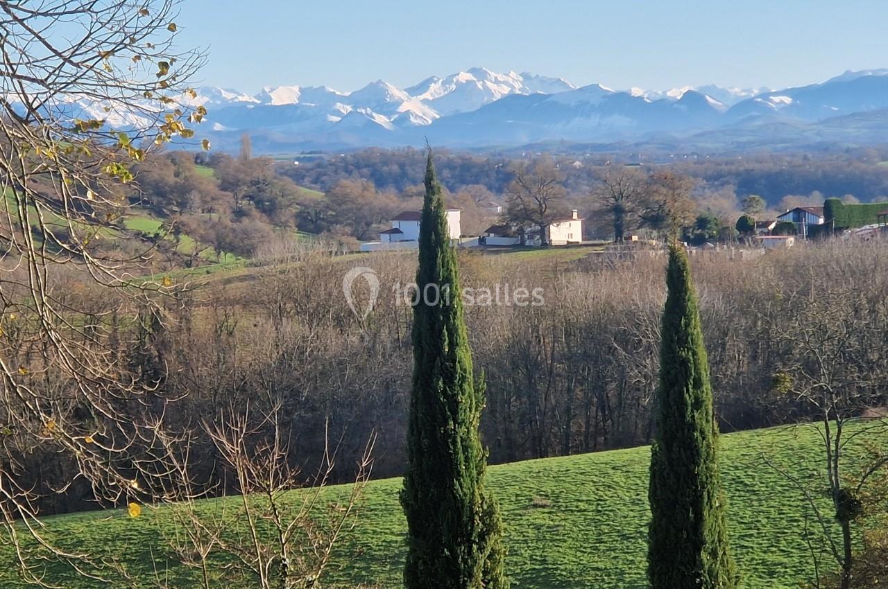 Paysage rural avec deux cyprès au premier plan, collines verdoyantes et montagnes enneigées à l'horizon.
