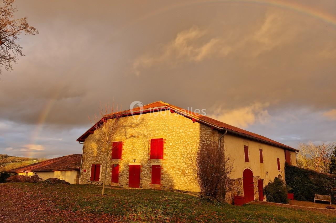 Maison en pierre avec volets rouges éclairée par le soleil couchant, sous un ciel nuageux avec un arc-en-ciel.