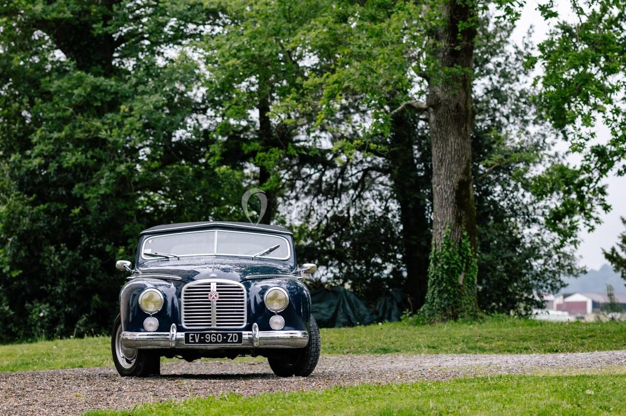 Voiture ancienne noire stationnée sur une allée gravillonnée, entourée d'arbres et de verdure.