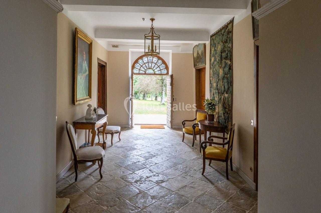 Couloir lumineux avec sol en pierre, mobilier ancien, tableaux aux murs et porte ouverte donnant sur un jardin verdoyant.