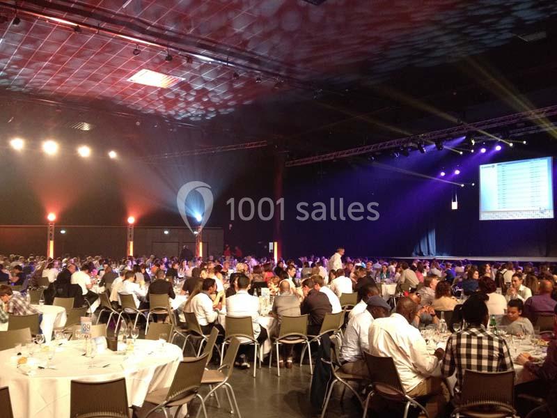 Salle de réception remplie de tables rondes avec des convives, éclairée par des projecteurs et un écran en fond.