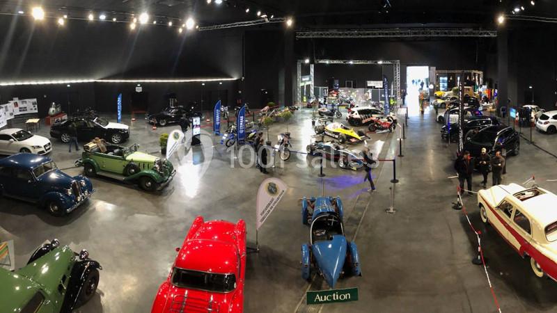 Vue d'une exposition de voitures classiques et modernes dans une grande salle éclairée, avec quelques visiteurs.