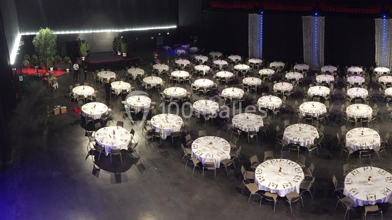 Salle de réception avec de nombreuses tables rondes dressées pour un événement, éclairage tamisé et décor sobre.