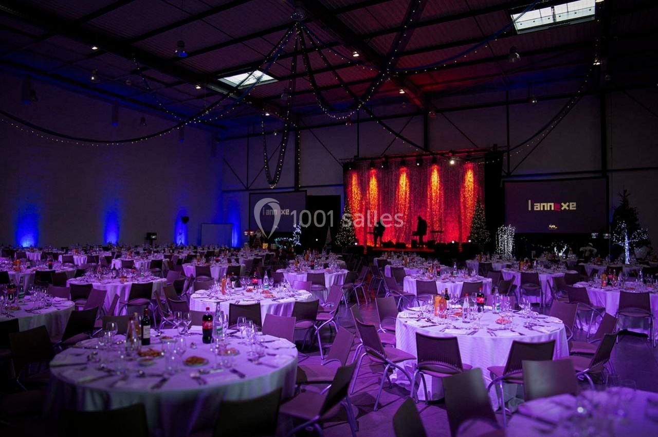 Salle de réception décorée avec des tables rondes dressées, éclairage tamisé et scène illuminée en arrière-plan.