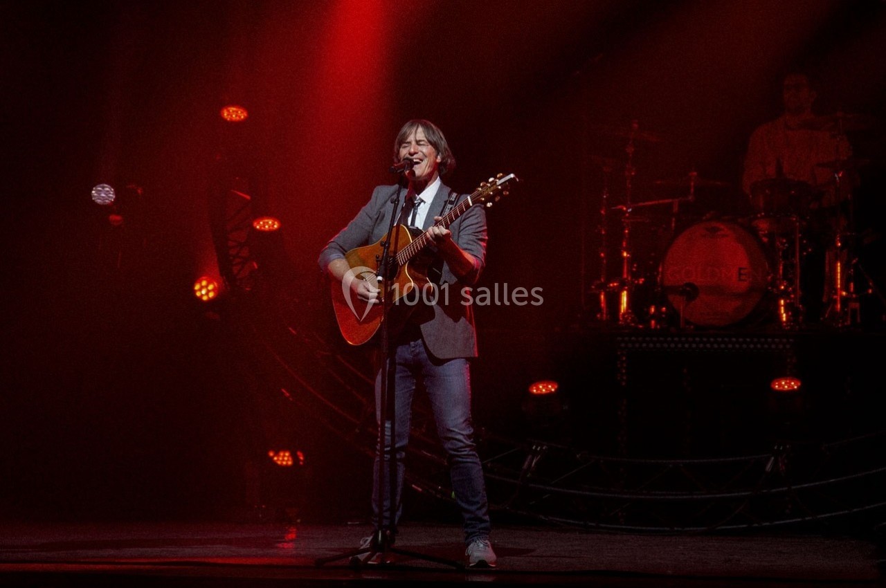Un musicien joue de la guitare sur une scène éclairée de lumières rouges, avec un batteur en arrière-plan.