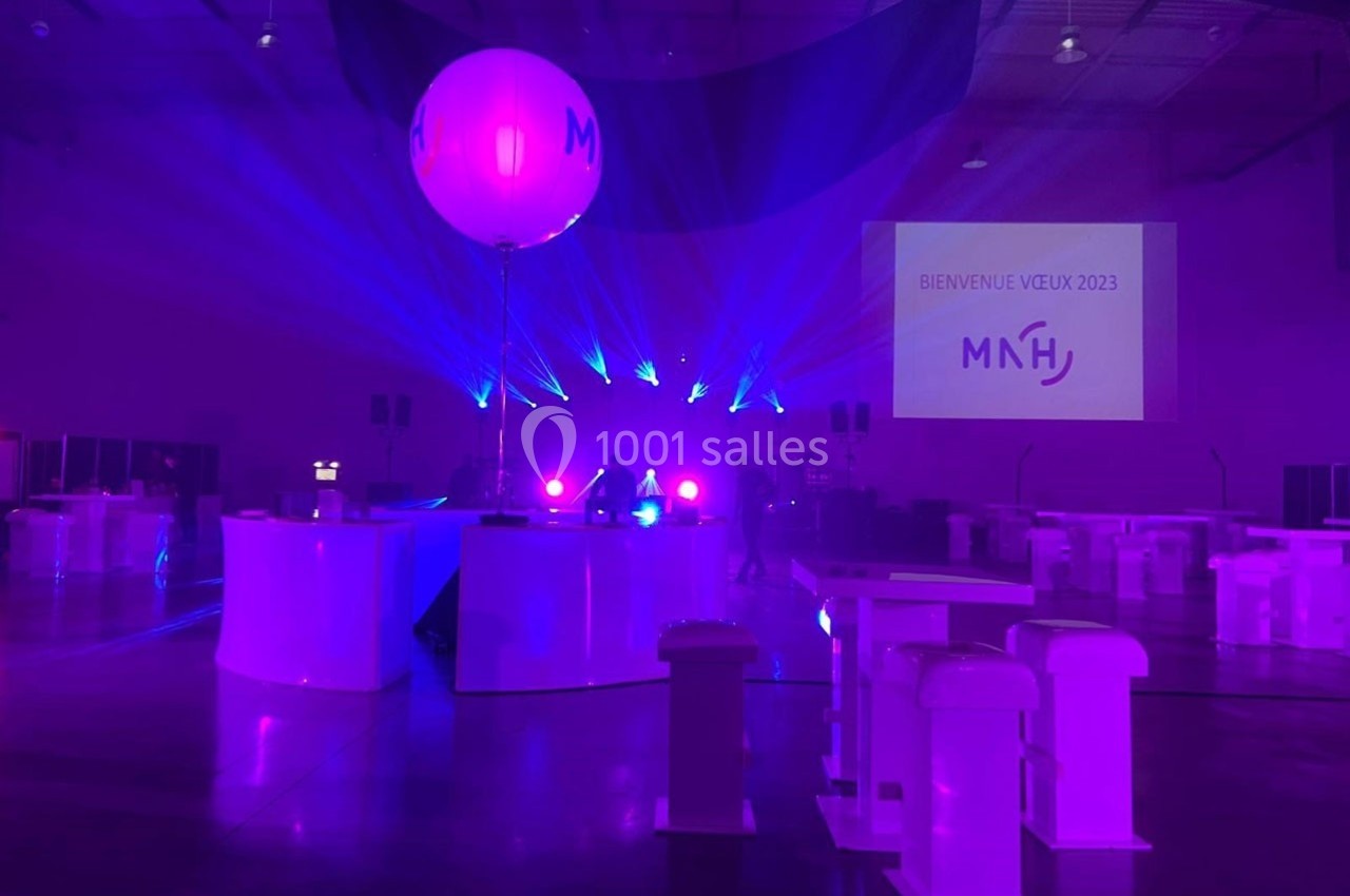 Salle événementielle éclairée par des lumières violettes, avec un ballon marqué ’MNH’ et un écran affichant ’Bienvenue Velix…