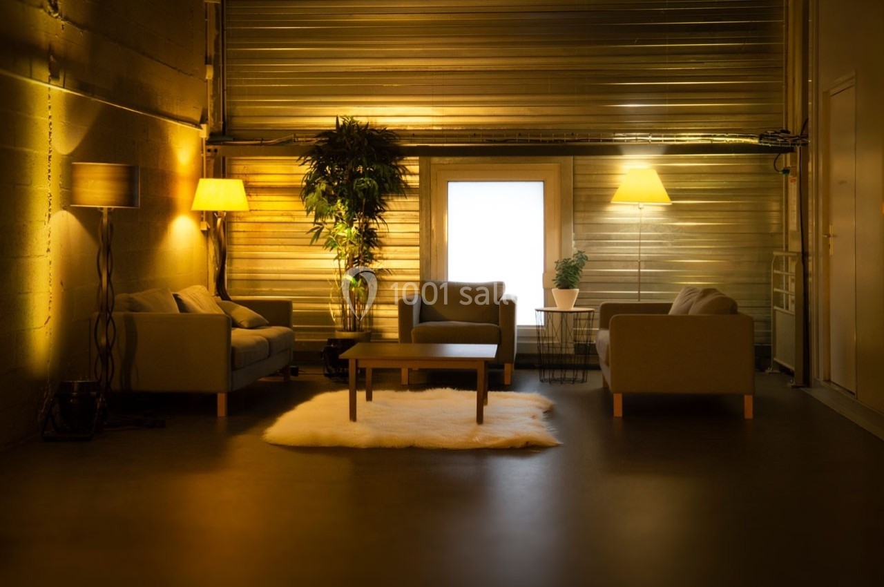 Espace salon chaleureux avec canapés, lampes et plantes, éclairé par une lumière douce devant une grande fenêtre.