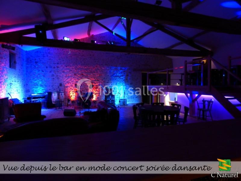 Salle de concert avec éclairage coloré, vue depuis un bar, équipée de chaises, tables et instruments de musique.