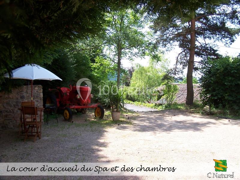 Cour extérieure avec un tracteur rouge, parasol blanc et végétation environnante, près d'un bâtiment en pierre.
