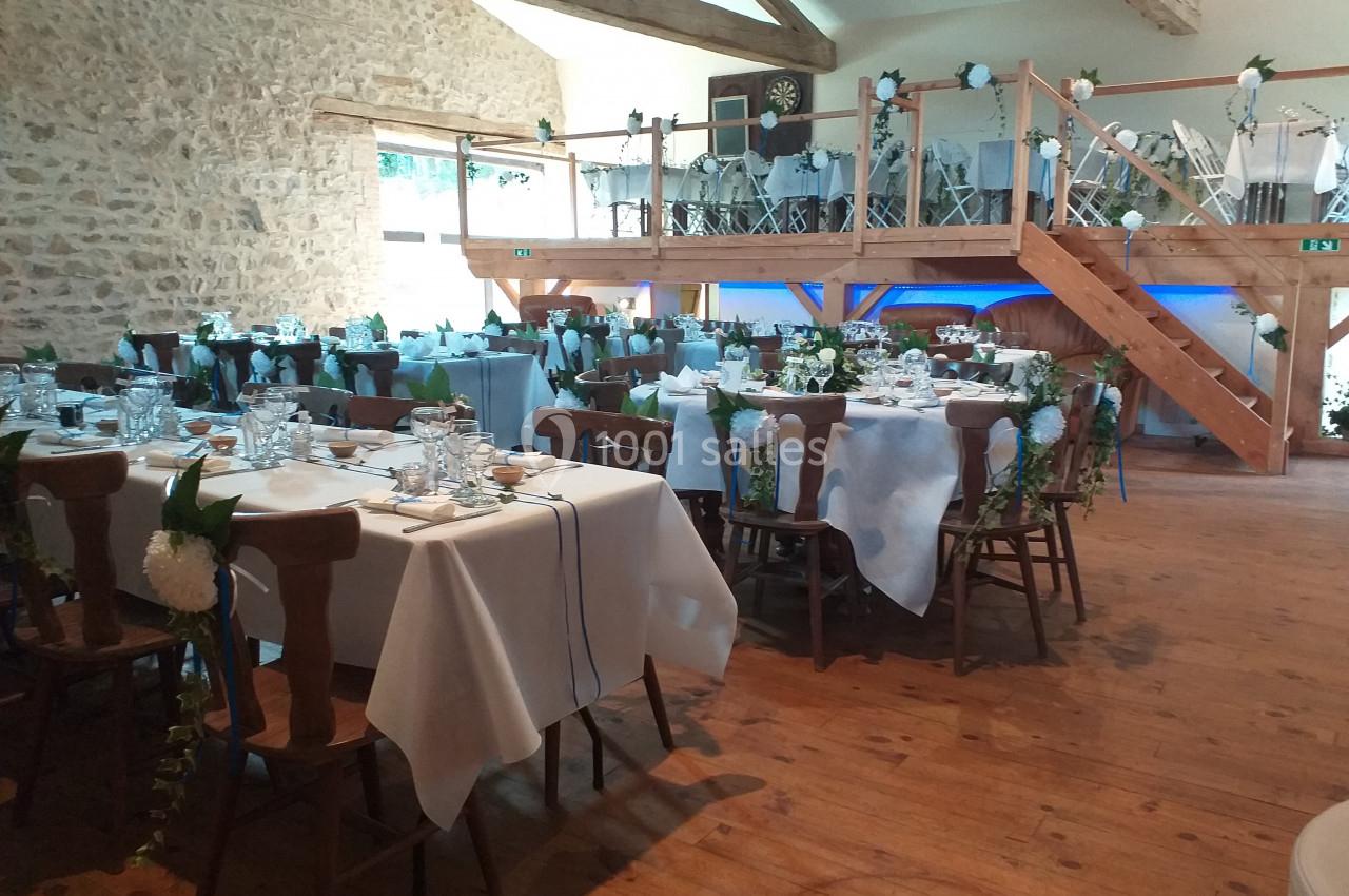 Salle de réception décorée pour un événement, avec tables dressées, chaises ornées et mezzanine en bois visible.