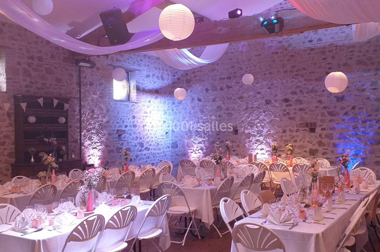 Salle en pierre décorée pour un événement, avec tables dressées, chaises blanches et lanternes suspendues.