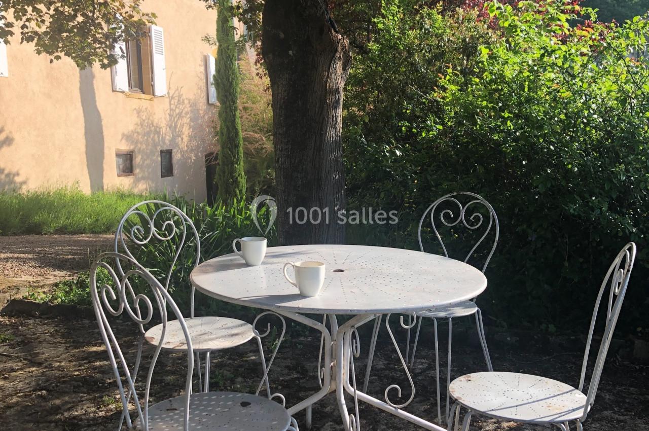 Table et chaises en métal blanc sous un arbre, avec deux tasses posées sur la table, dans un jardin ensoleillé.