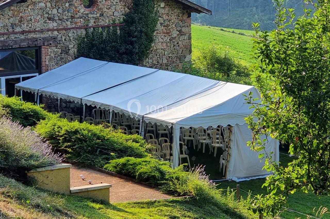 Tente blanche installée à côté d'une maison en pierre, entourée de verdure et de chaises visibles à l'intérieur.