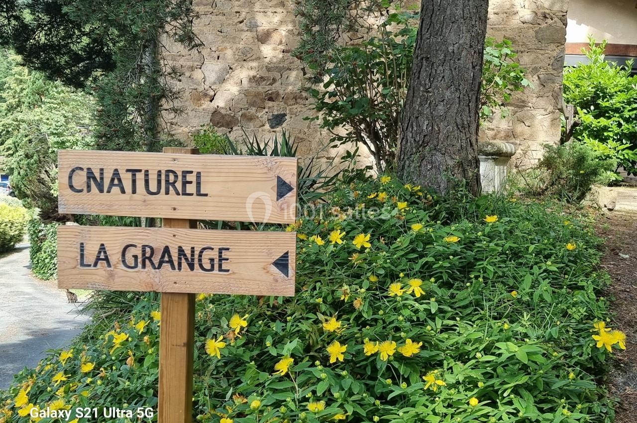 Panneaux en bois indiquant ’Cnaturel’ et ’La Grange’, entourés de fleurs jaunes et de végétation près d'un mur en pierre.
