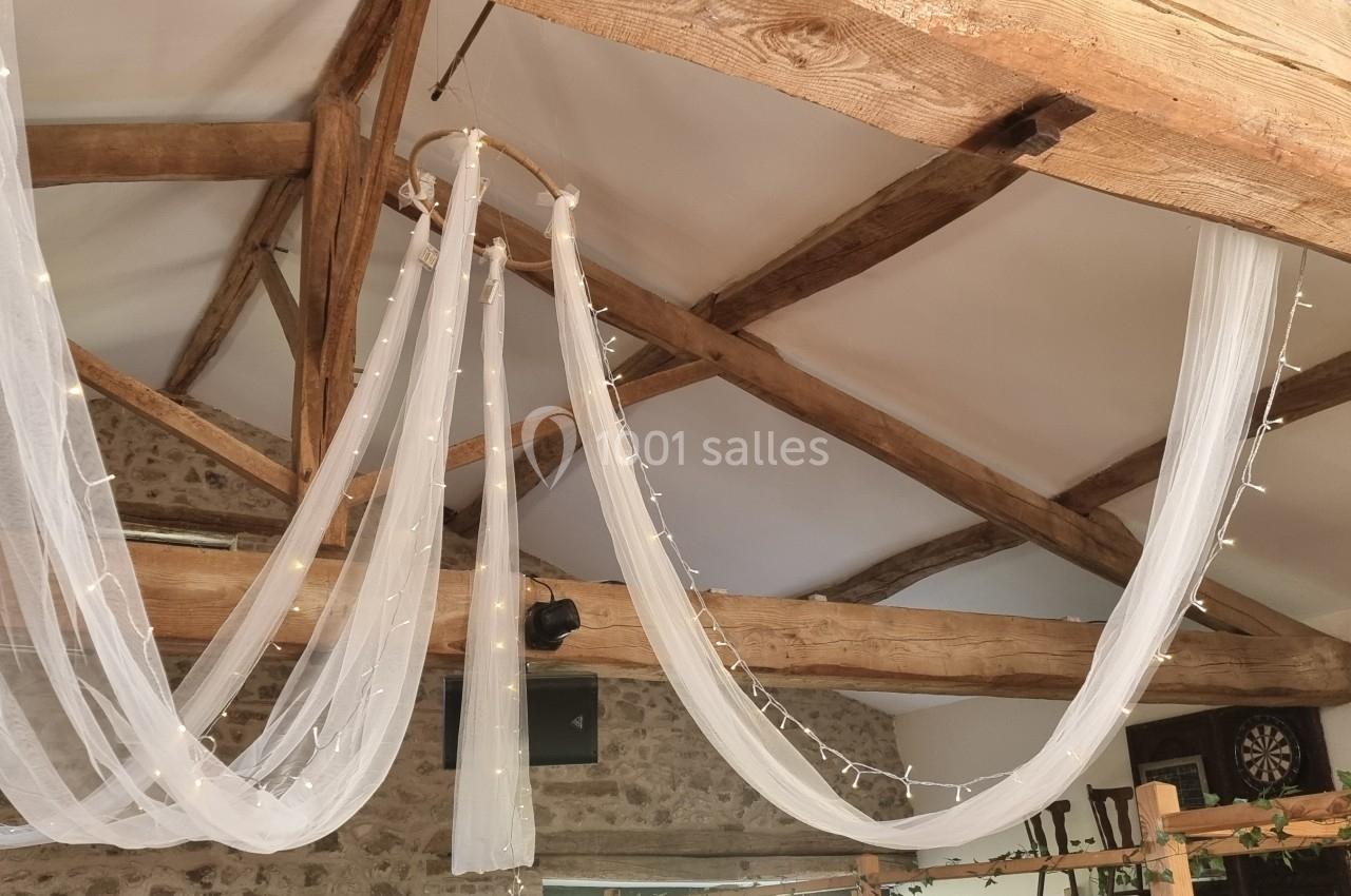 Plafond avec poutres en bois apparentes décorées de voilages blancs et de guirlandes lumineuses.