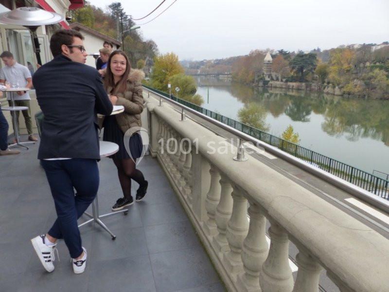 Deux personnes discutent debout sur une terrasse surplombant une rivière et un paysage arboré en automne.