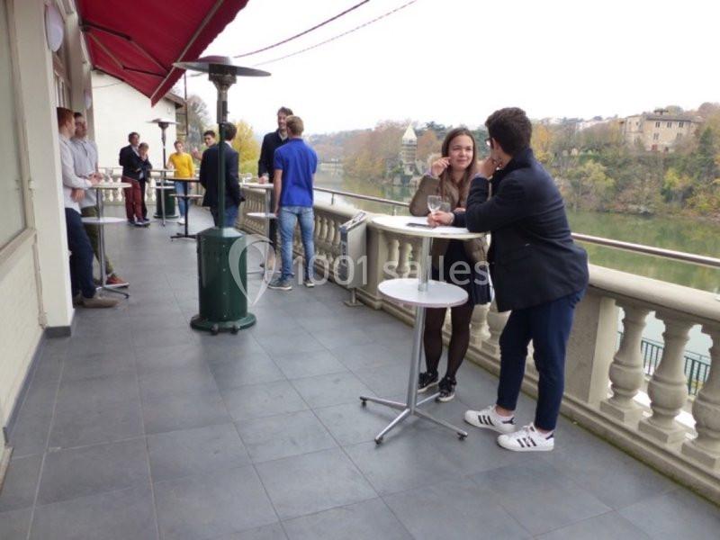 Des personnes discutent debout autour de tables hautes sur une terrasse avec vue sur un fleuve et des arbres.