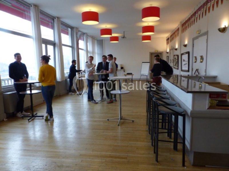 Personnes discutant en petits groupes dans une salle lumineuse avec des tables hautes et un bar en arrière-plan.