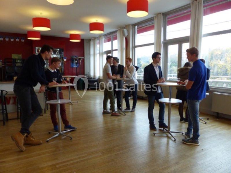 Personnes discutant debout autour de tables hautes dans une salle lumineuse avec des fenêtres et des lampes rouges.