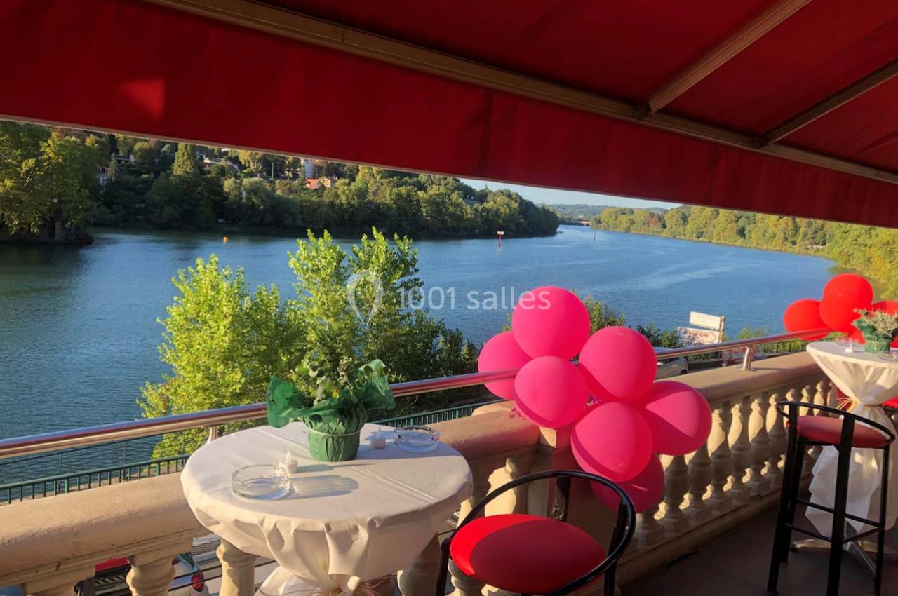 Vue d'une terrasse avec tables dressées, ballons roses et panorama sur une rivière bordée de verdure.