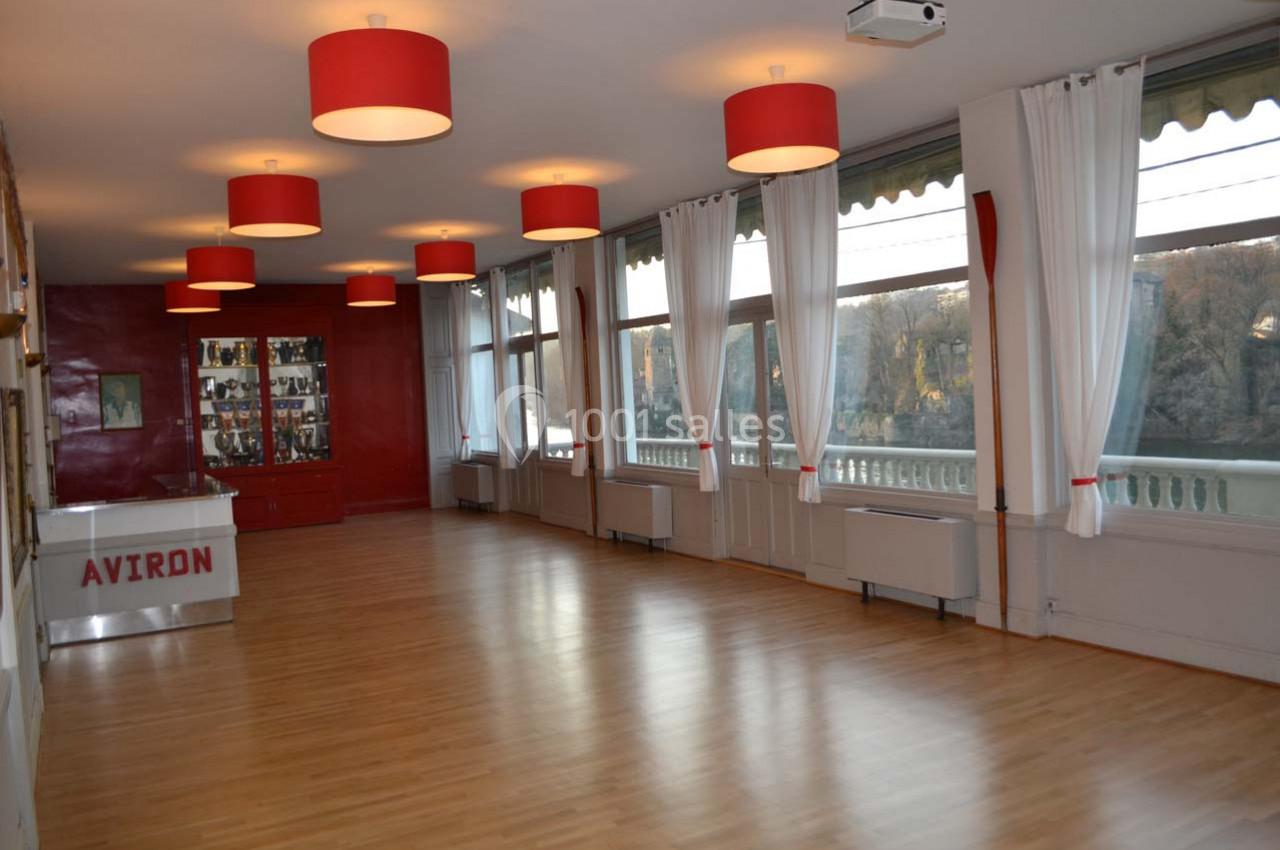 Salle lumineuse avec parquet, grandes fenêtres donnant sur l'extérieur, rideaux blancs et luminaires rouges.