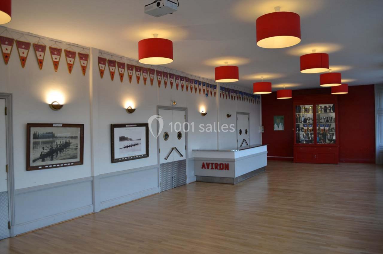 Salle lumineuse avec parquet, murs décorés de photos encadrées, fanions suspendus et comptoir marqué ’Avron’.