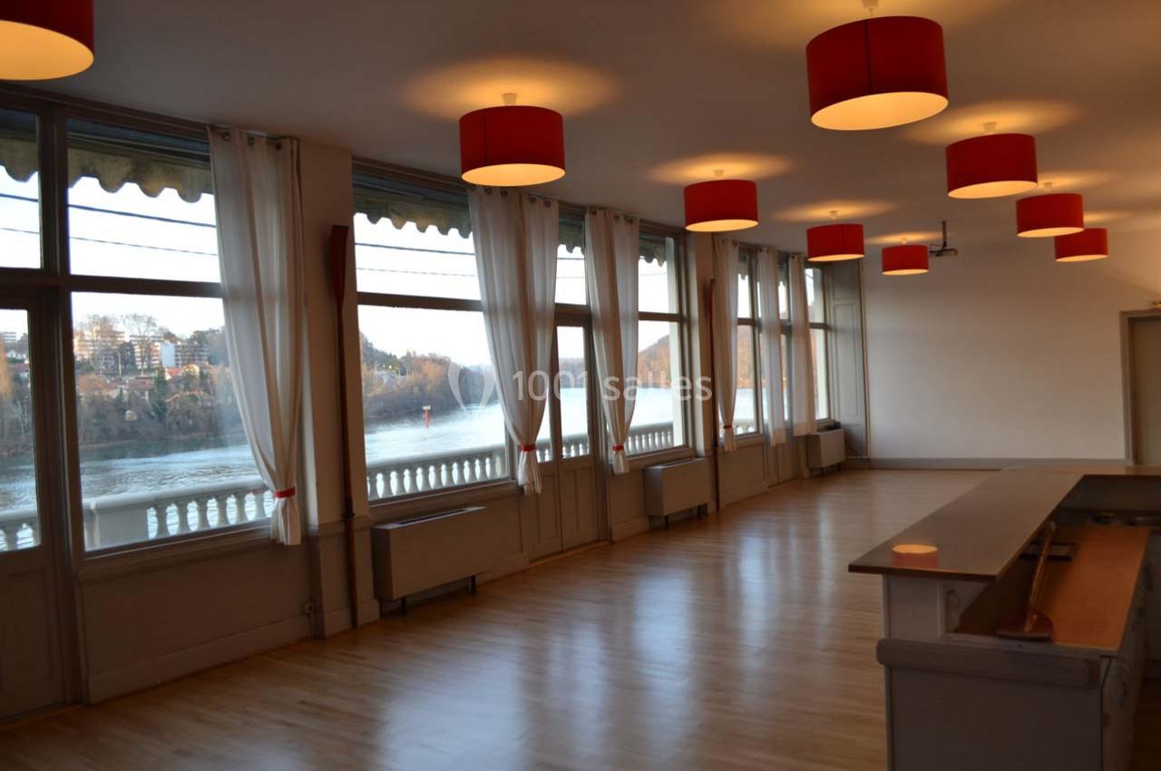 Salle lumineuse avec parquet en bois, grandes baies vitrées donnant sur une rivière et plafonniers rouges.