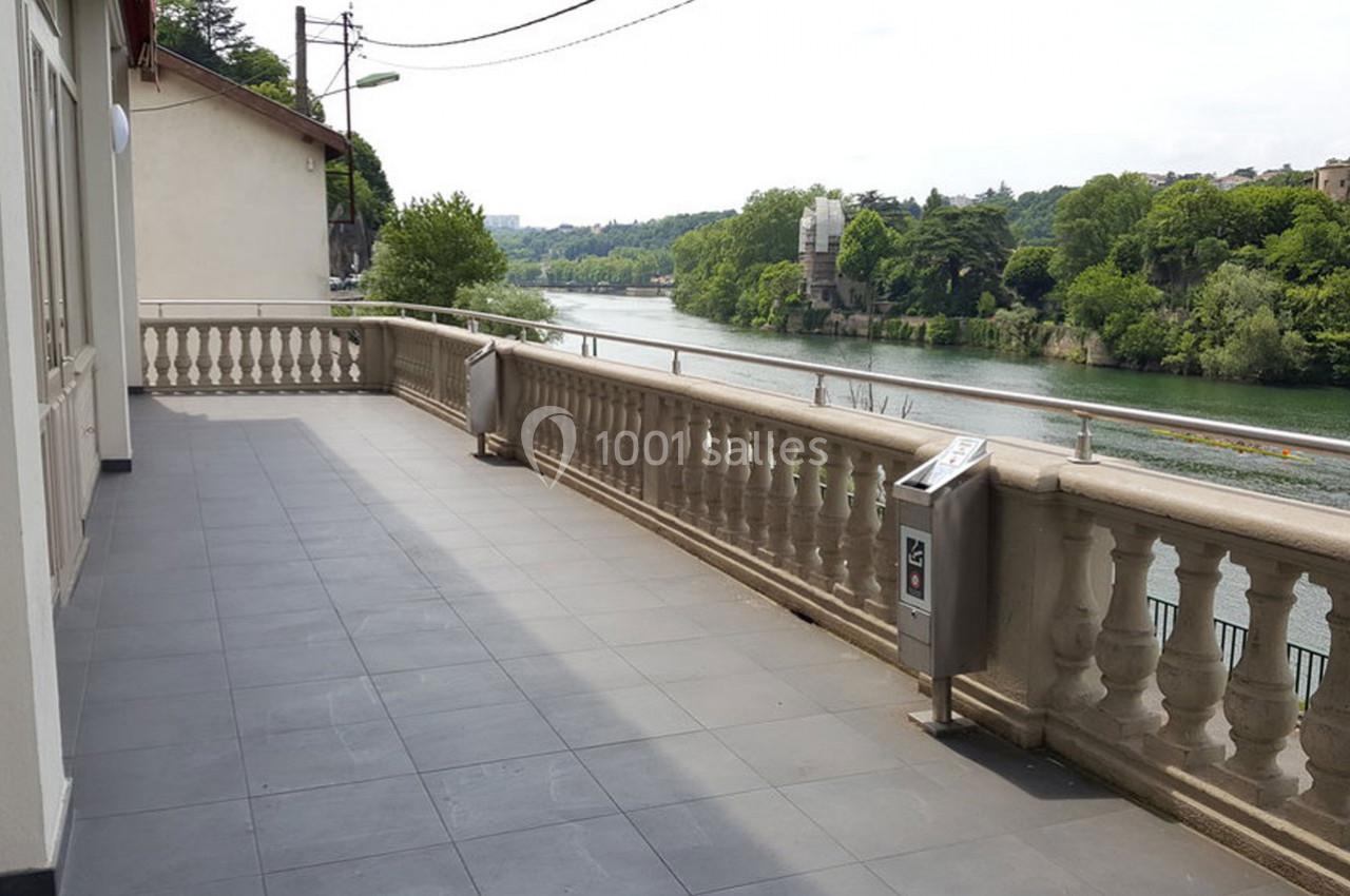 Terrasse carrelée avec balustrade donnant sur une rivière bordée de végétation et bâtiments en arrière-plan.