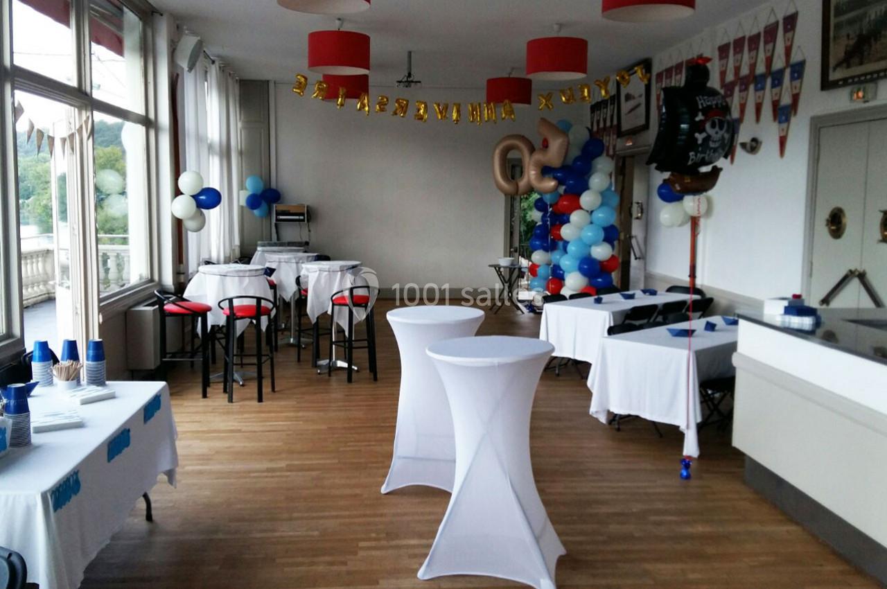 Salle décorée pour une fête d'anniversaire avec ballons, guirlande et tables hautes recouvertes de nappes blanches.
