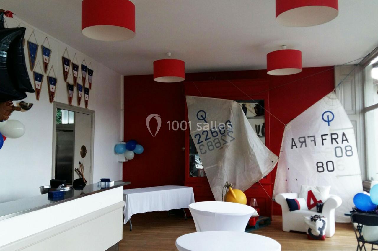 Salle décorée avec des voiles de bateau, des fanions nautiques et des ballons, avec un comptoir et des tables blanches.