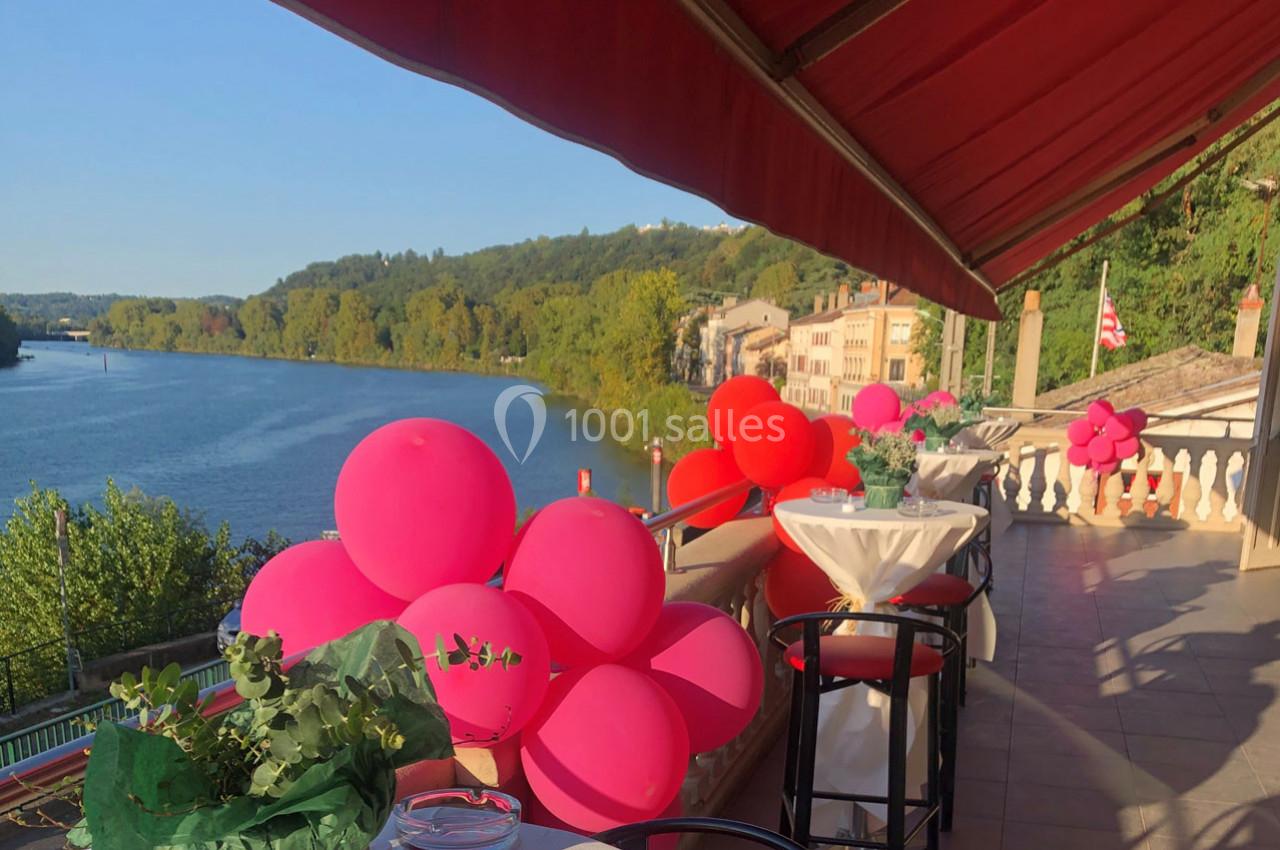 Terrasse avec vue sur une rivière bordée d'arbres, décorée de ballons roses et rouges sous un auvent rouge.