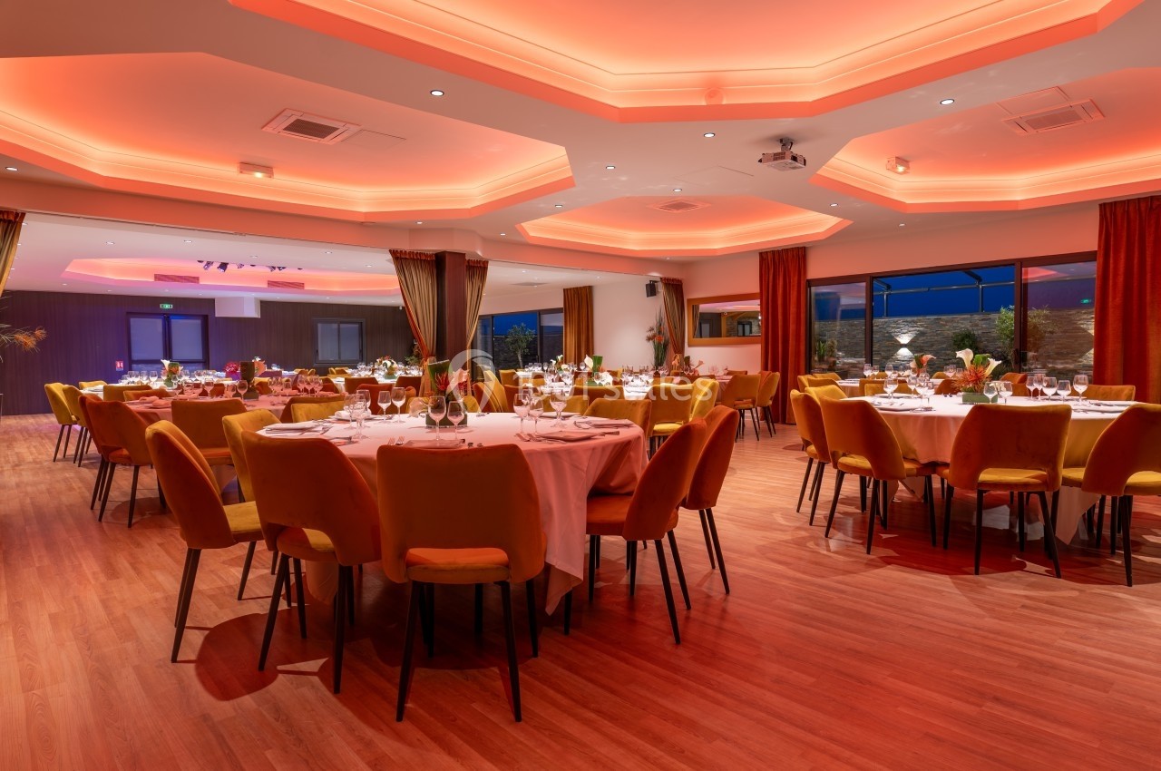 Salle de réception élégante avec tables rondes dressées, éclairage tamisé et grandes baies vitrées.