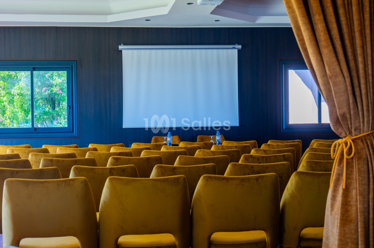 Salle de conférence avec des rangées de chaises jaunes, un écran de projection blanc et des fenêtres donnant sur l'extérieur.