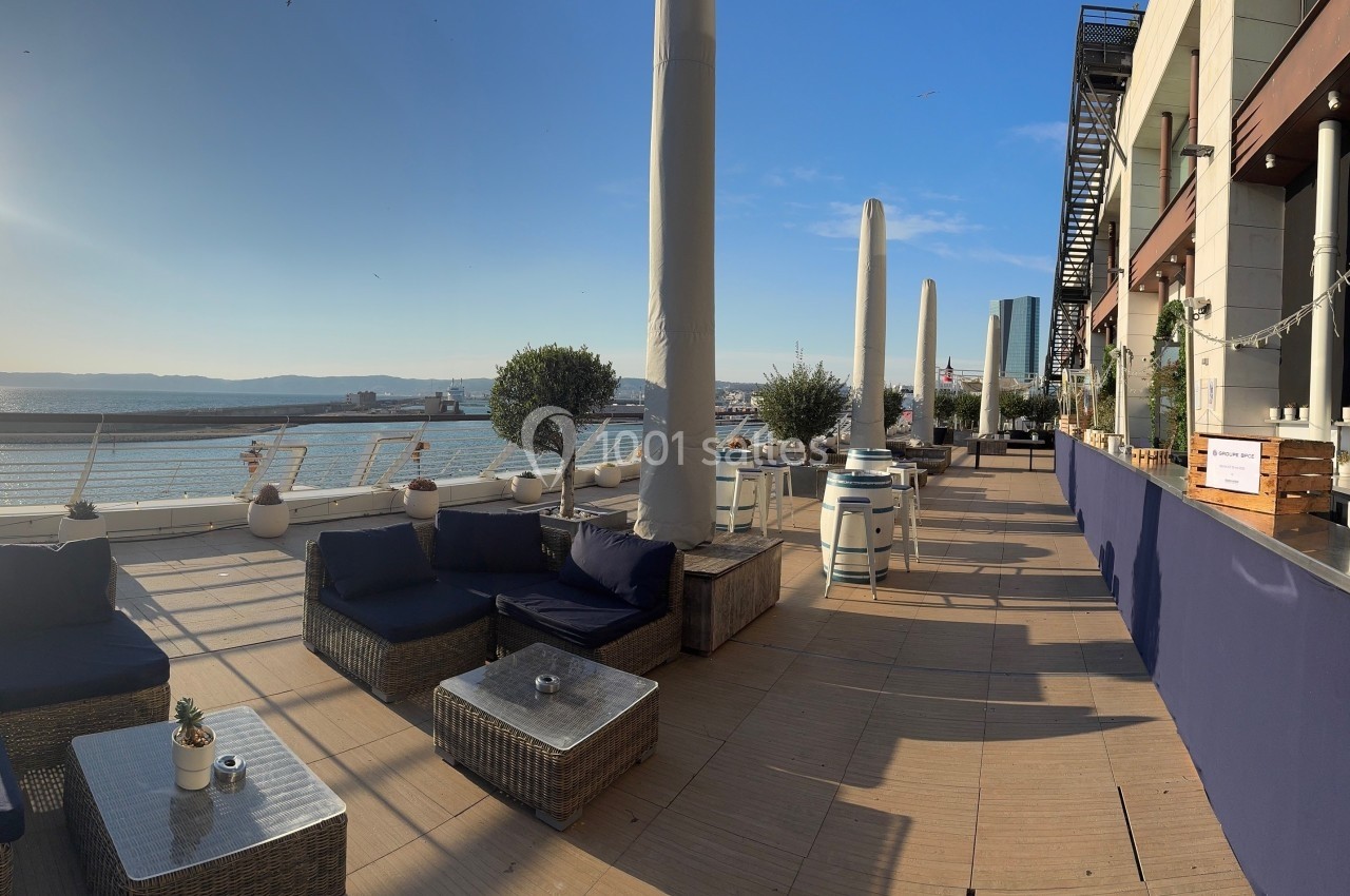 Terrasse ensoleillée avec mobilier en rotin, vue sur la mer et colonnes blanches, bordée par un bâtiment moderne.