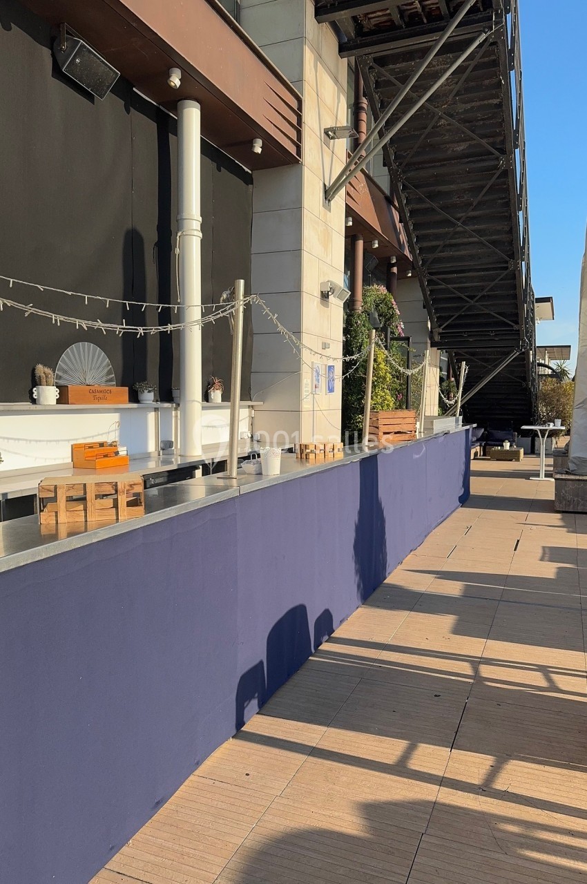 Comptoir extérieur vide avec nappe bleue, éclairage suspendu et terrasse ensoleillée près d'une structure métallique.