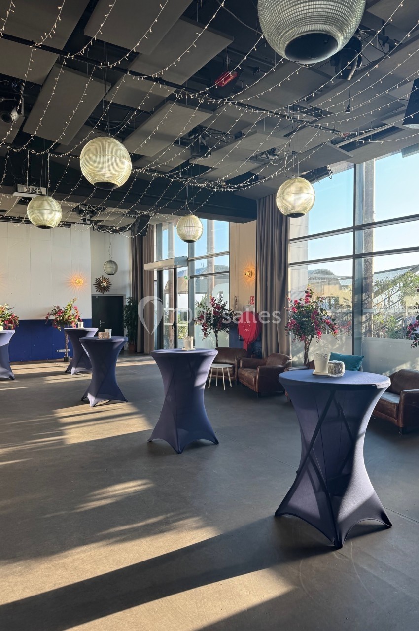 Salle lumineuse décorée de guirlandes lumineuses, tables hautes drapées de bleu et arrangements floraux colorés.
