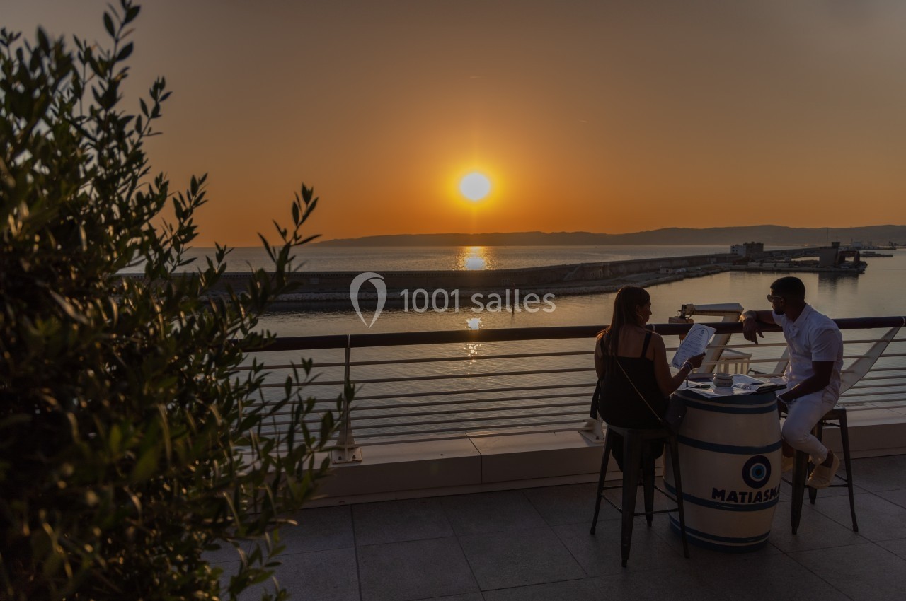 Un couple dîne en terrasse face à un coucher de soleil sur la mer, avec une vue sur un port au loin.