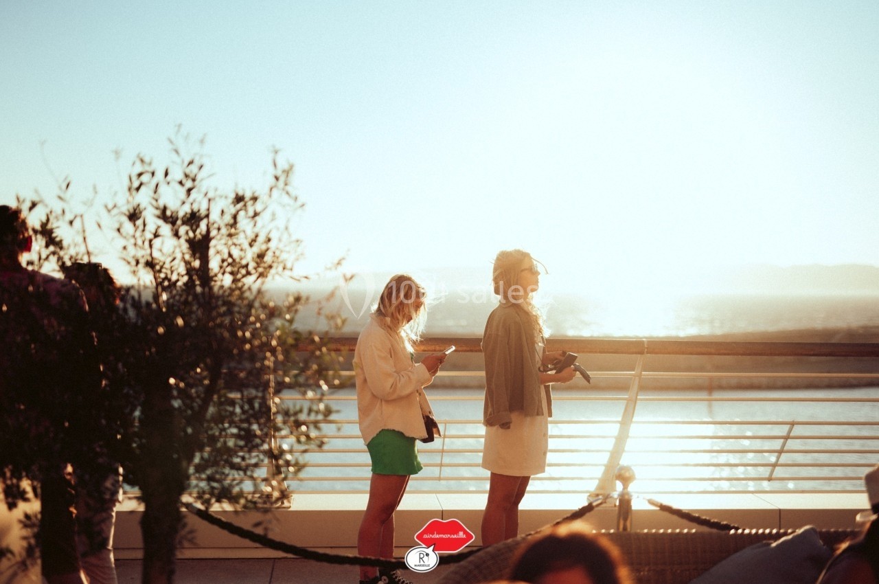 Deux femmes debout sur une terrasse ensoleillée avec vue sur la mer, l'une regardant son téléphone.