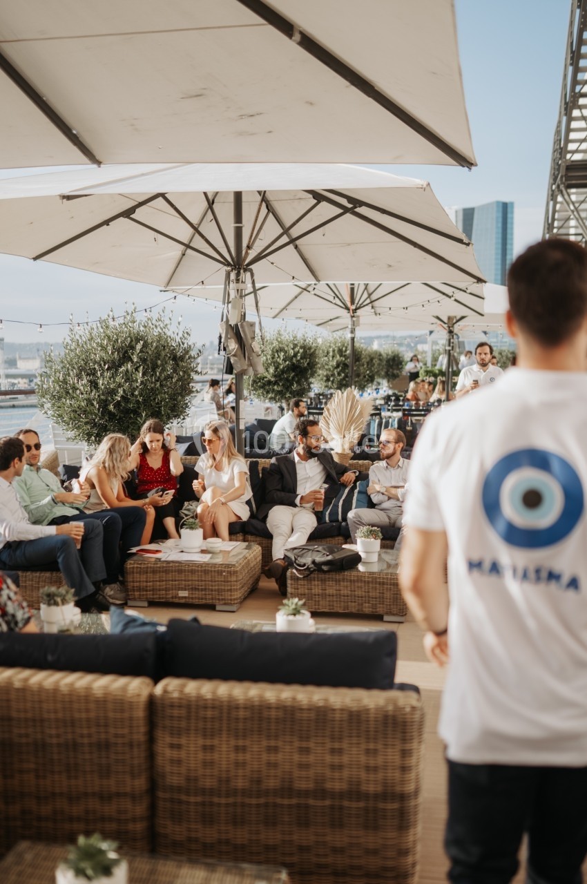 Des personnes discutent et se détendent sur une terrasse aménagée avec des parasols et des canapés en osier.
