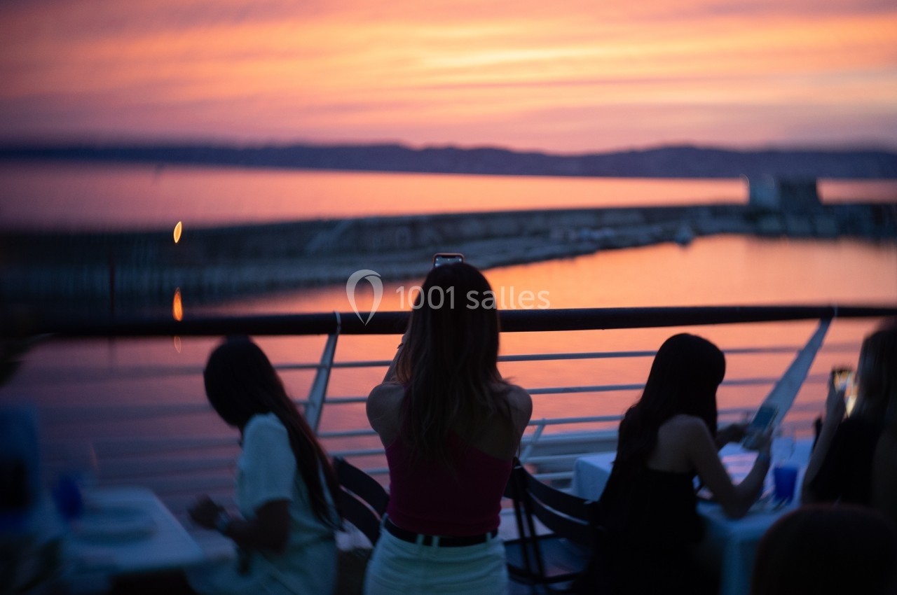 Une femme prend une photo d'un coucher de soleil depuis une terrasse surplombant un plan d'eau.