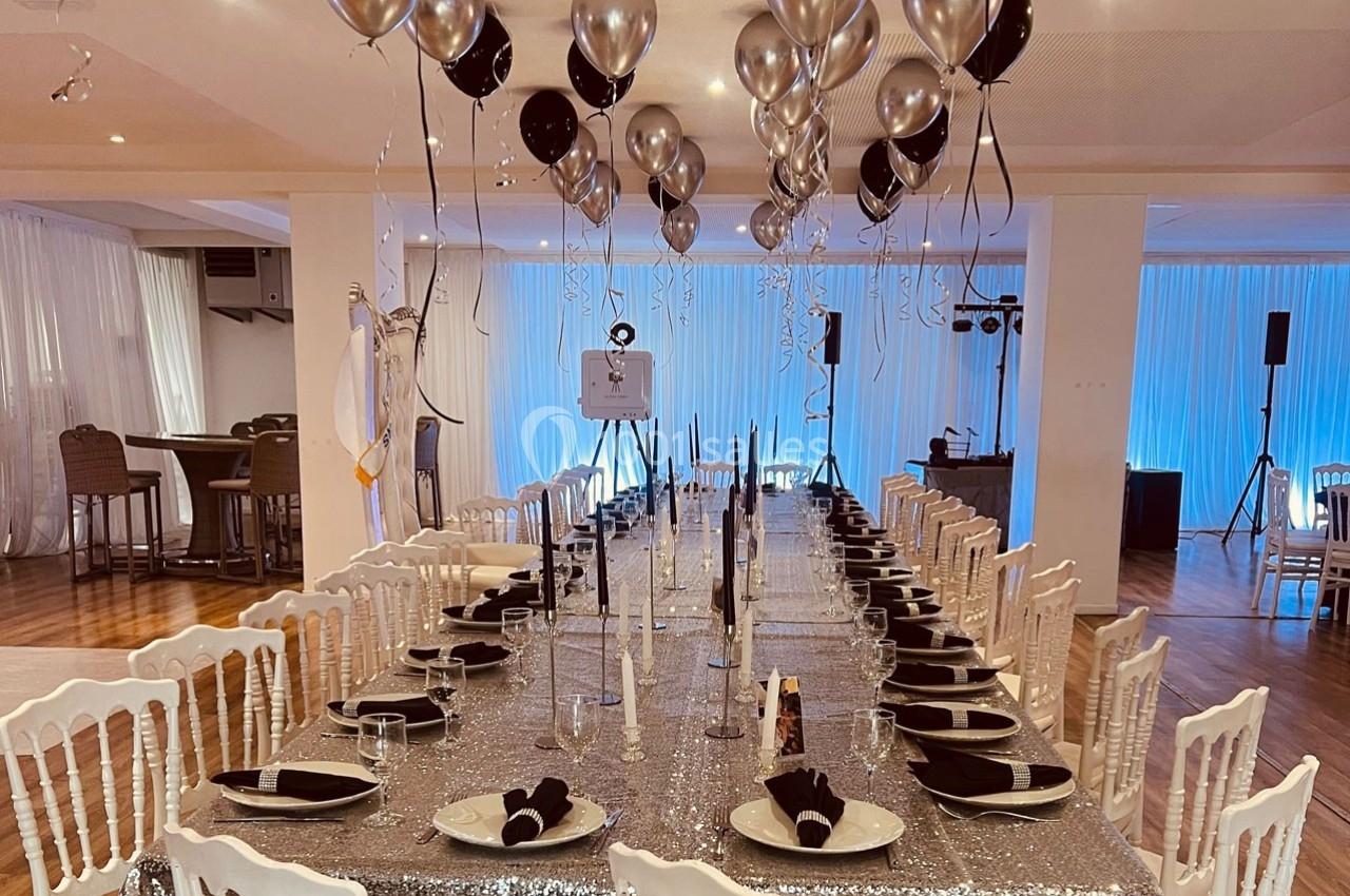 Grande table rectangulaire décorée pour un événement, avec nappes scintillantes, ballons argentés et noirs, et vaisselle…