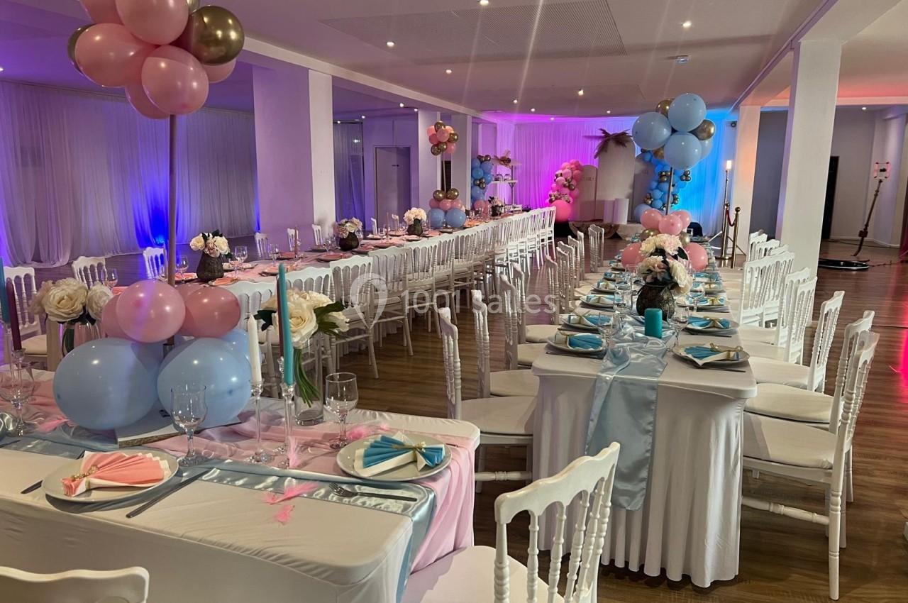 Salle décorée pour une réception avec tables alignées, nappes roses et bleues, ballons et compositions florales.