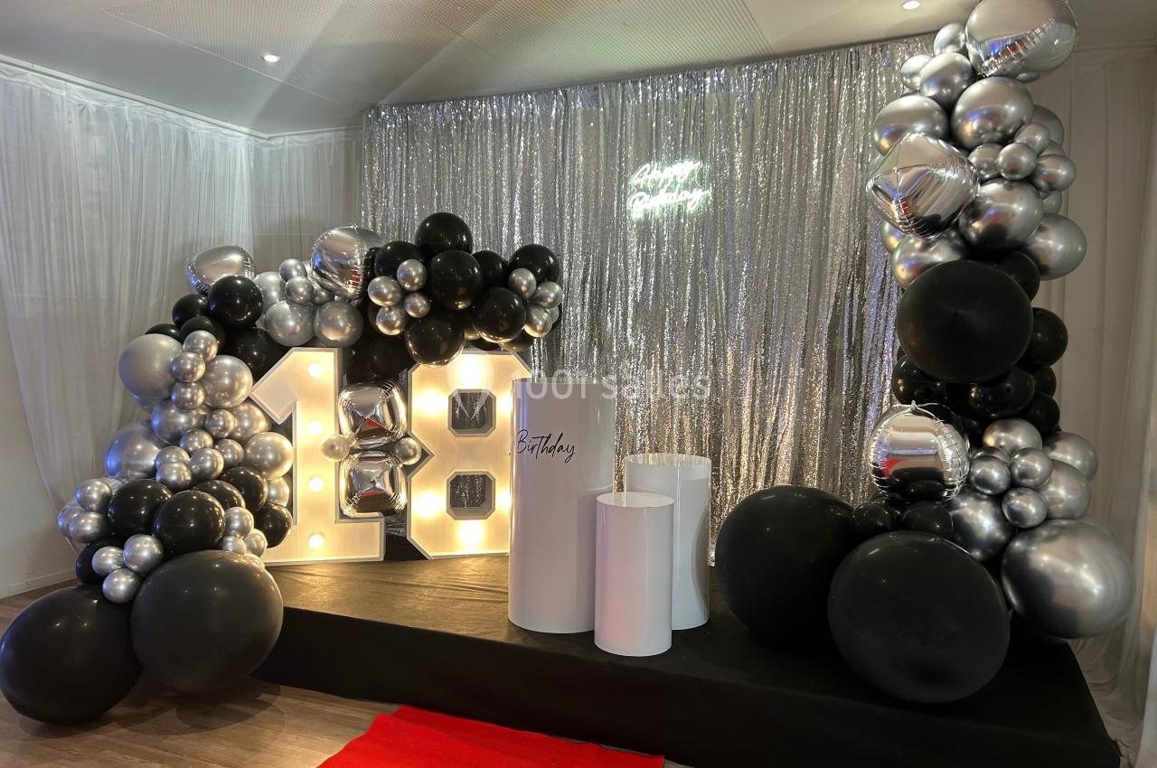 Décoration de fête avec ballons noirs et argentés, rideau scintillant, chiffres lumineux ’18’ et tapis rouge.