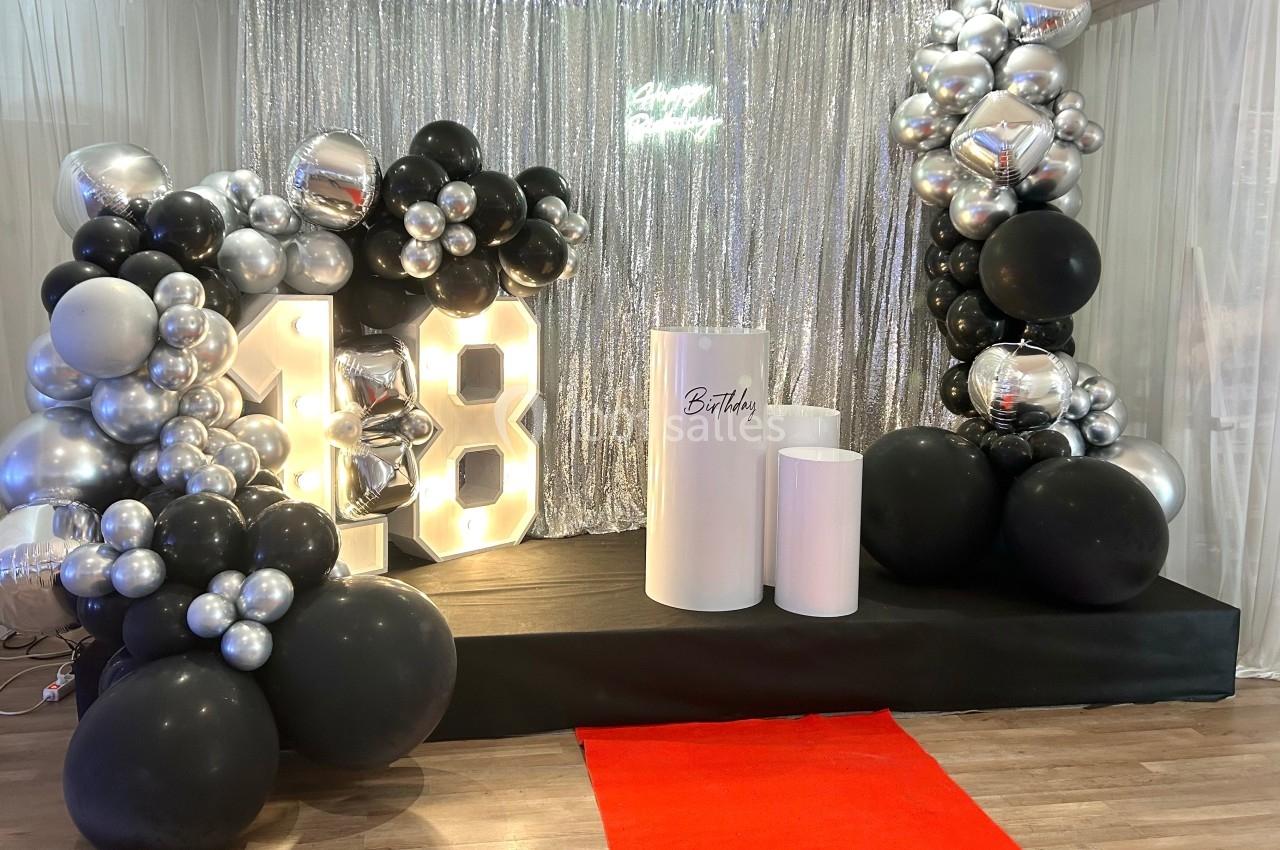 Décor de fête avec ballons noirs et argentés, chiffres lumineux ’18’, rideau scintillant et tapis rouge.