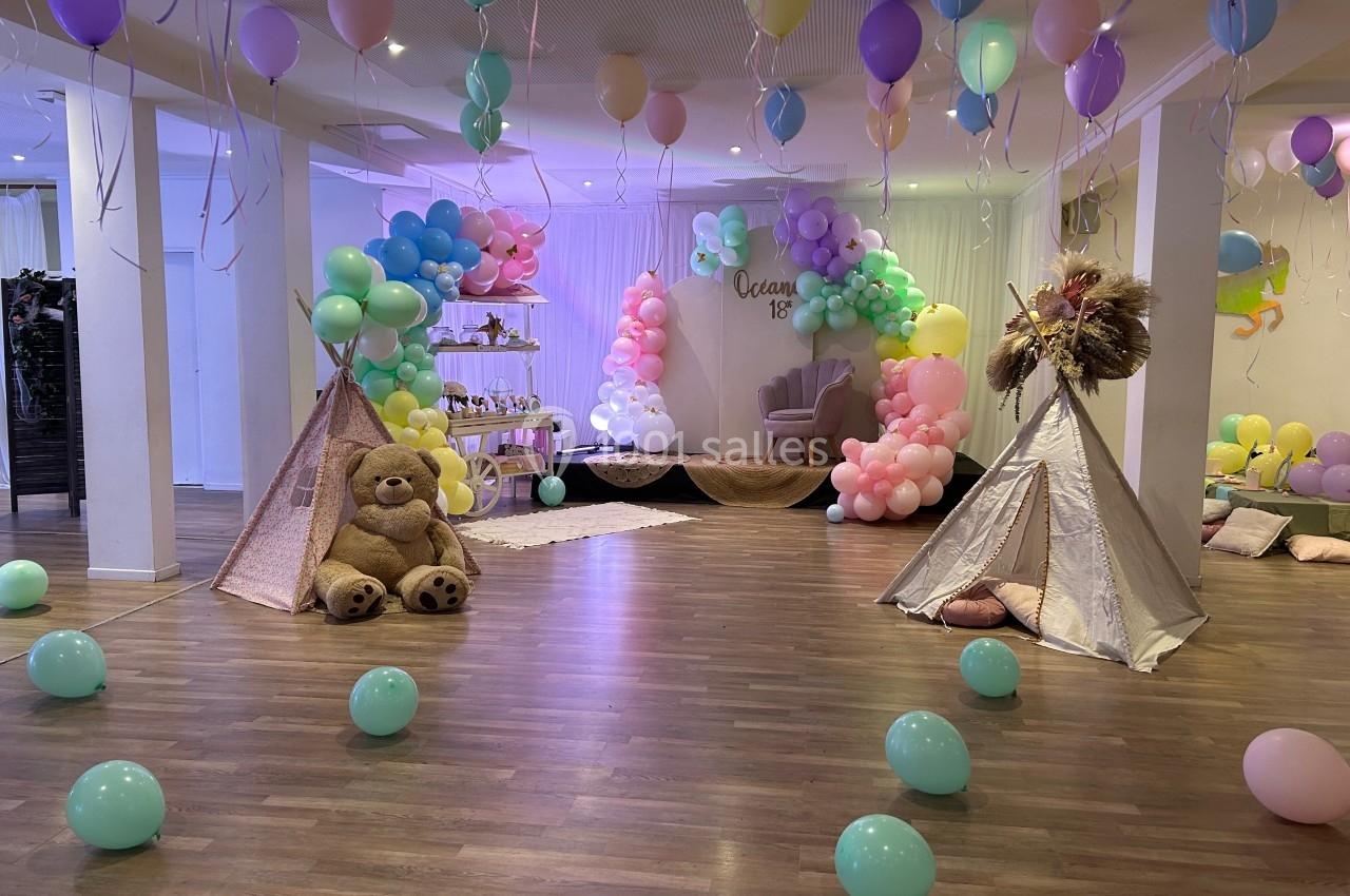 Salle décorée pour une fête avec ballons colorés, tipis, un grand ours en peluche et une arche de ballons pastel.