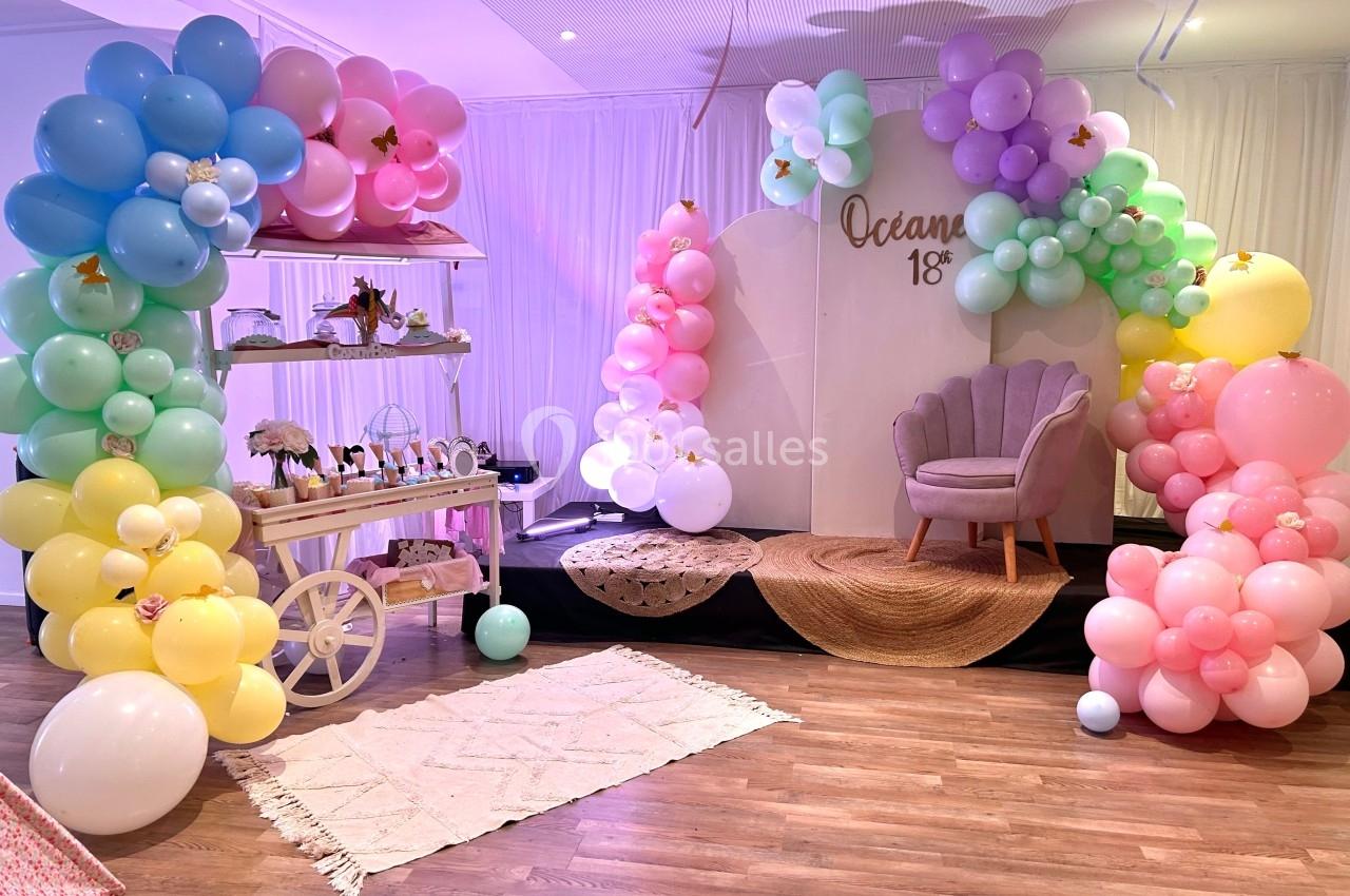 Décoration festive avec arche de ballons pastel, fauteuil rose et chariot décoratif sur un sol en bois.