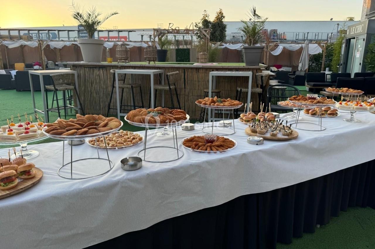 Buffet en extérieur avec une table garnie de viennoiseries, sandwichs, amuse-bouches et verrines, au coucher du soleil.